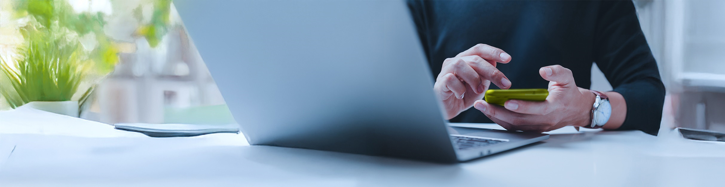 Person sitzt vor Laptop mit Smartphone in der Hand und tippt darauf