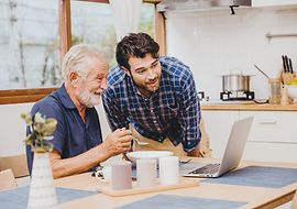 Junger Mann schaut mit älterem Mann, der einen Laptop bedient, auf den Laptop und freuen sich