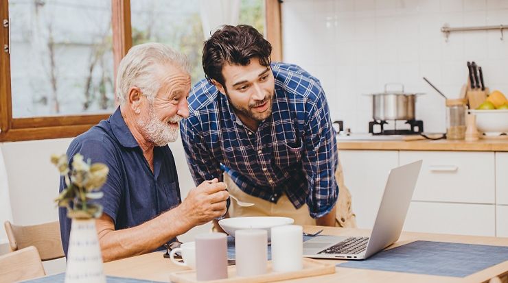 Junger Mann schaut mit älterem Mann, der einen Laptop bedient, auf den Laptop und freuen sich