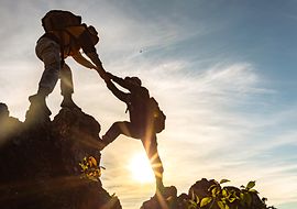 Zwei Wandernde reichen sich die Hände, um einen großen Abstand zwischen zwei Felsen zu überwinden