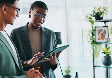 Zwei Personen in Business-Outfits im Gespräch, eine Person hält ein Tablet.