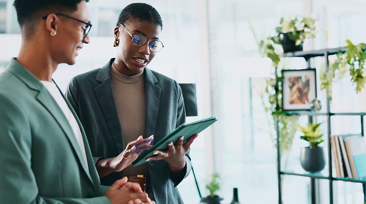 Zwei Personen in Business-Outfits im Gespräch, eine Person hält ein Tablet.