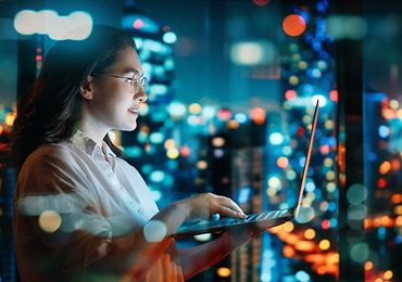 Frau arbeitet nachts am Laptop, im Hintergrund Skyline