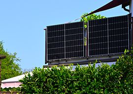 Solarpanele auf einem Balkongeländer mit grüner Hecke und blauem Himmel im Hintergrund.