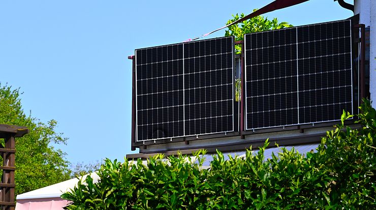 Solarpanele auf einem Balkongeländer mit grüner Hecke und blauem Himmel im Hintergrund.