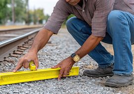 Person misst Schotterbett an Bahngleisen mit einer Wasserwaage und einem Maßband.