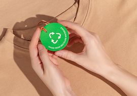 Zwei Hände zeigen ein Nachhaltigkeitssiegel, das an einem T-Shirt befestigt ist, in die Kamera