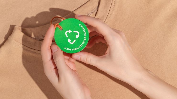 Zwei Hände zeigen ein Nachhaltigkeitssiegel, das an einem T-Shirt befestigt ist, in die Kamera
