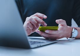 Person sitzt vor Laptop mit Smartphone in der Hand und tippt darauf