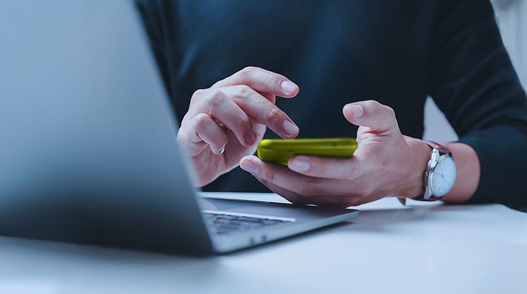 Person sitzt vor Laptop mit Smartphone in der Hand und tippt darauf