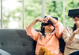 Child and adult playing with VR headsets on a sofa in front of a window.