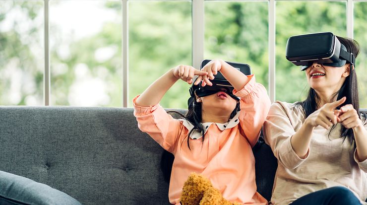 Child and adult playing with VR headsets on a sofa in front of a window.