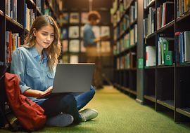 Eine Frau sitzt am Boden einer Bibliothek mit Laptop, Bücherregale im Hintergrund.