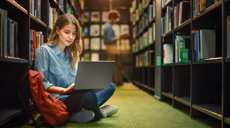 Eine Frau sitzt am Boden einer Bibliothek mit Laptop, Bücherregale im Hintergrund.