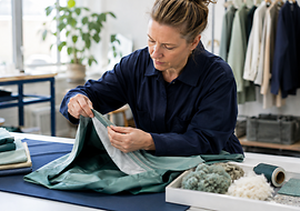 Eine Frau prüft sorgfältig ein Stoffstück in einem Atelier, umgeben von Textilien und Materialien.