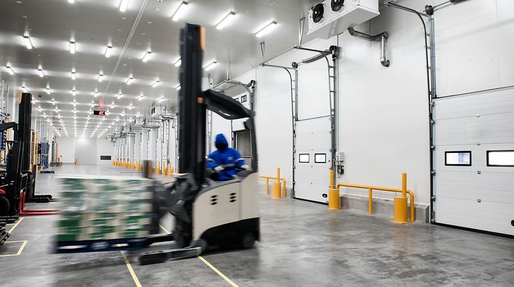 Person hebt mithilfe eines Gabelstaplers eine Palette Kisten in einer Lagerhalle