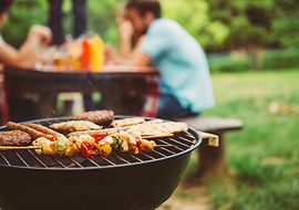 Im Vordergrund: Grill mit Fleisch und Gemüse. Im Hintergrund: Menschen sitzen an einem Tisch im Garten.