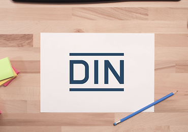 Blatt Papier mit DIN Logo auf einem Schreibtisch. Daneben ein Bleistift und Post-Its.