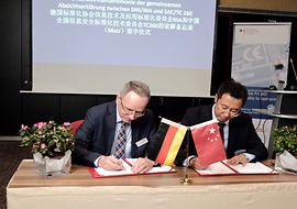 Volker Jacumeit und Mr. Liu Xiangang unterschreiben eine Vereinbarung. Eien deutsche und chinesiche Flagge stehen auf dem Tisch.
