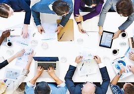 Meeting table from above with many people at the edge and numerous pieces of paper on the table