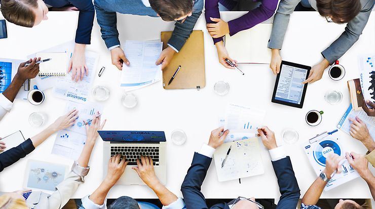 Meeting table from above with many people at the edge and numerous pieces of paper on the table