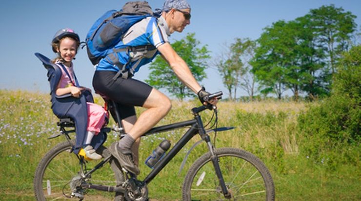 Person mit Kind auf einem Fahrrad auf einem Weg in ländlicher Umgebung unterwegs.