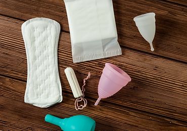 Verschiedene Menstruationsprodukte auf Holzuntergrund, inklusive Binden und Menstruationstassen.